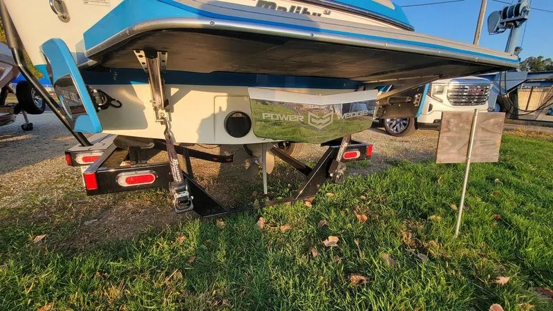 Slide: The Image of 2015 Malibu 24 MXZ boat rear view, parked on grass with trailer supports visible. - 26