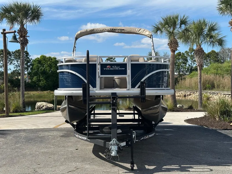 Slide: The Image of 2025 Sun Tracker Fishin' Barge 20 DLX pontoon boat on trailer, surrounded by palm trees. - 2