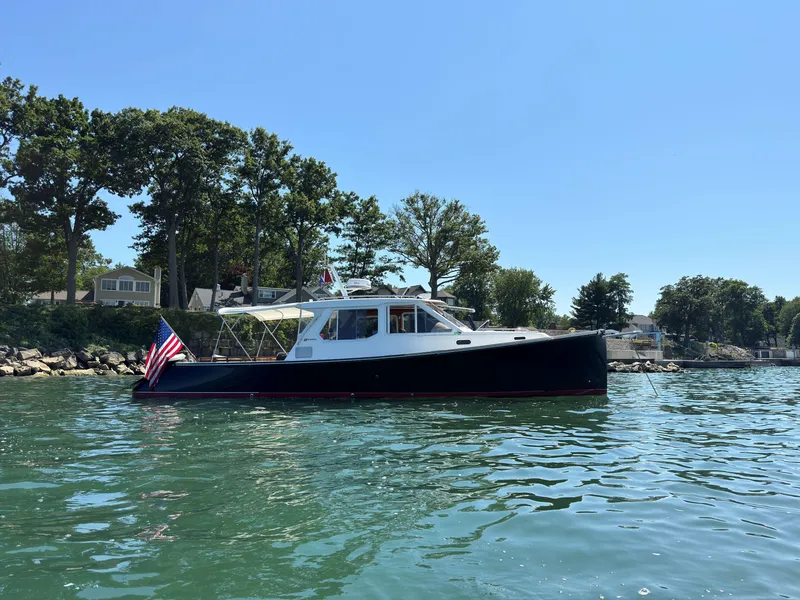 Slide: The Image of 2005 True North 38 Heritage boat on calm water, with trees and houses in the background. - 1