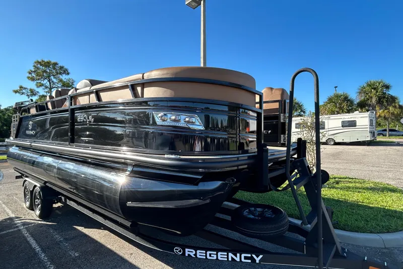 The Image of 2025 Regency 230 LE3 Sport pontoon boat on trailer, parked outdoors under clear blue sky. - 0