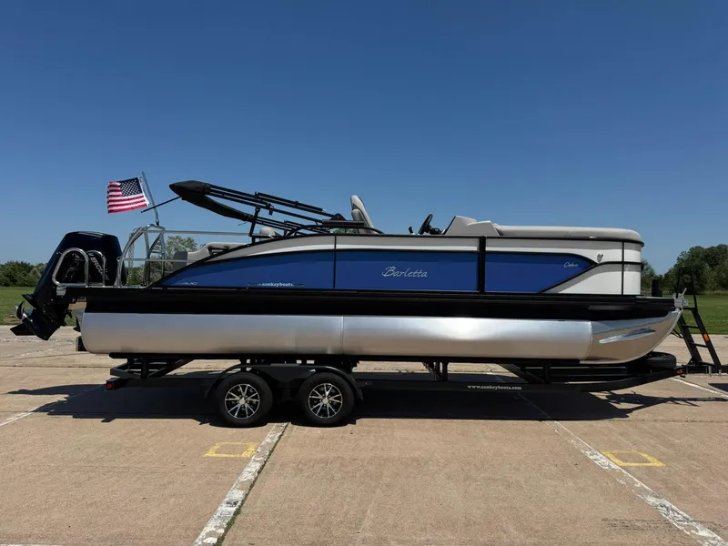 Slide: The Image of 2025 Barletta Cabrio 22UC pontoon boat on trailer, American flag, clear sky. - 12