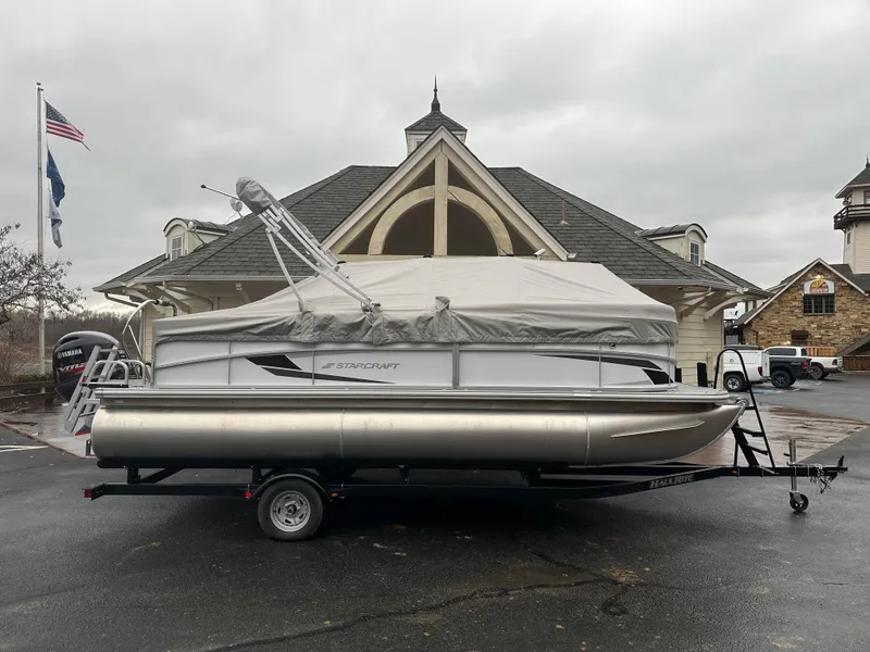 Slide: The Image of 2024 Starcraft LX 20 CB pontoon boat on trailer, parked outside a building on a cloudy day. - 6