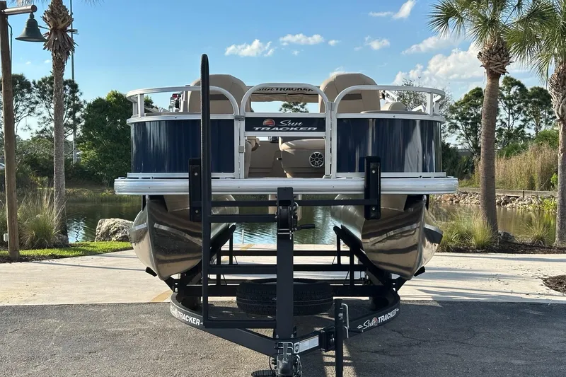 Slide: The Image of 2025 Sun Tracker Bass Buggy 18 DLX pontoon boat on trailer, parked near a scenic lake. - 2