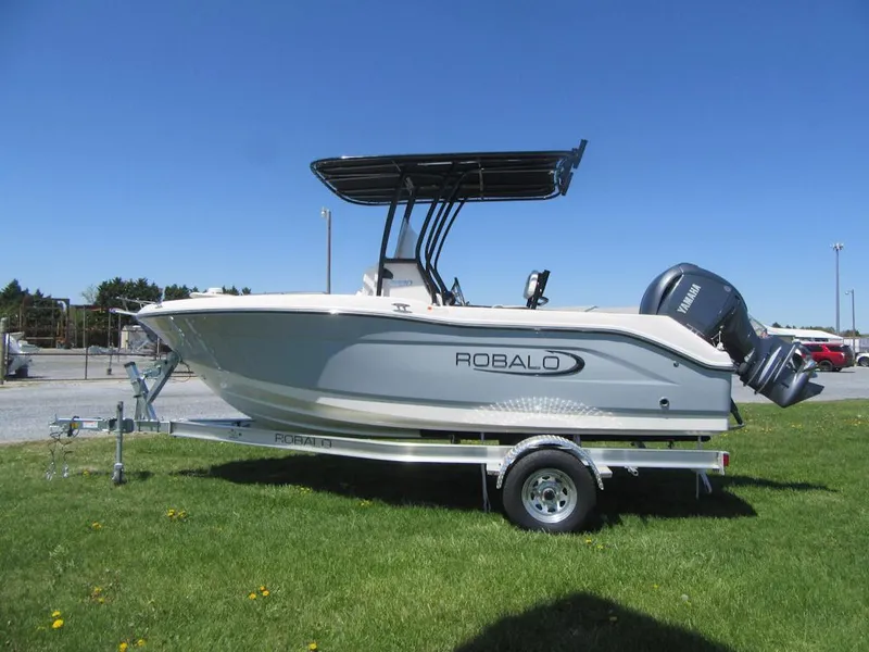 Slide: The Image of 2025 Robalo R180 Center Console boat on trailer, parked on grass under clear blue sky. - 2