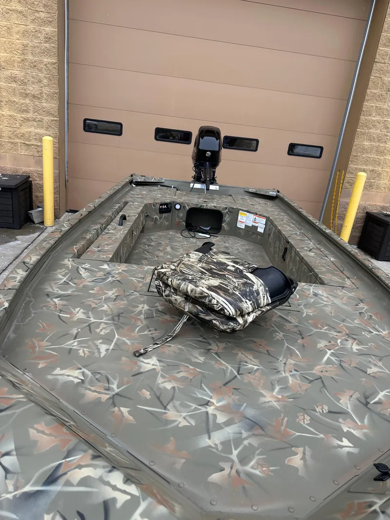 Slide: The Image of 2025 Tracker Grizzly 1654 T Sportsman boat with camouflage design, parked near a garage door. - 2