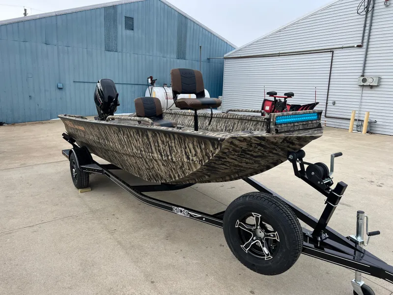 Slide: The Image of 2025 Excel 1651 River Warrior boat on trailer, camo design, parked near industrial buildings. - 1