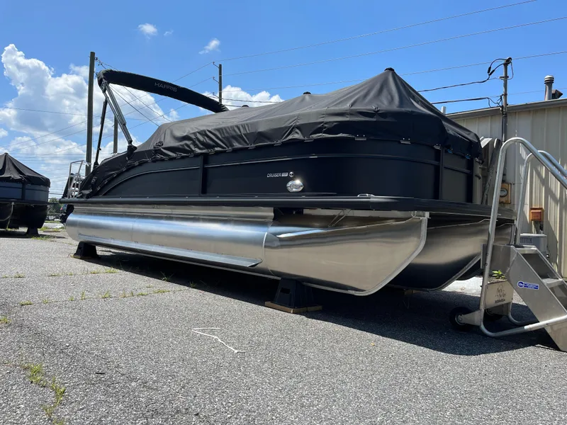 Slide: The Image of 2025 Harris Cruiser 230 pontoon boat with black cover, parked outdoors on a sunny day. - 3