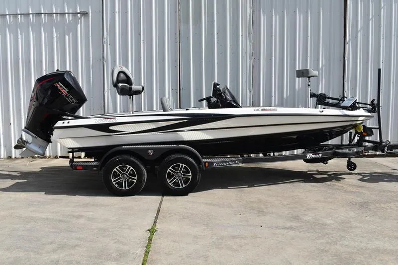 Slide: The Image of 2025 Triton 19 TrX Patriot boat on trailer, side view, parked by metal wall. - 4