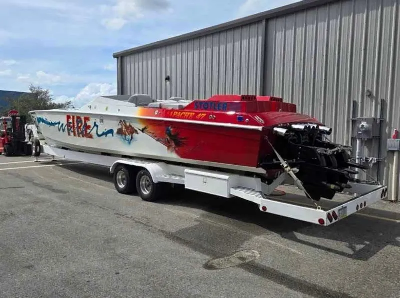 Slide: The Image of 1994 Apache 47 Fire Water speedboat on trailer, vibrant graphics, parked near metal building. - 2