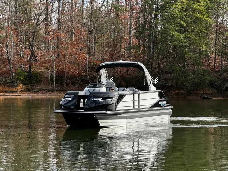 Slide: The Image of 2025 Montara Surf Boss 2.0 25 pontoon boat on a calm lake, surrounded by trees. - 2