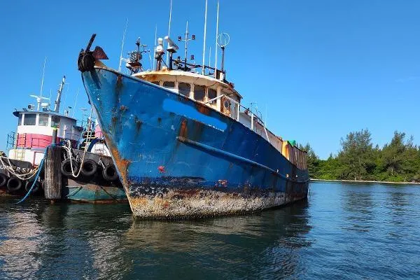 Slide: The Image of 1983 commercial freezer trawler docked, blue hull, calm water, clear sky, trees in background. - 1