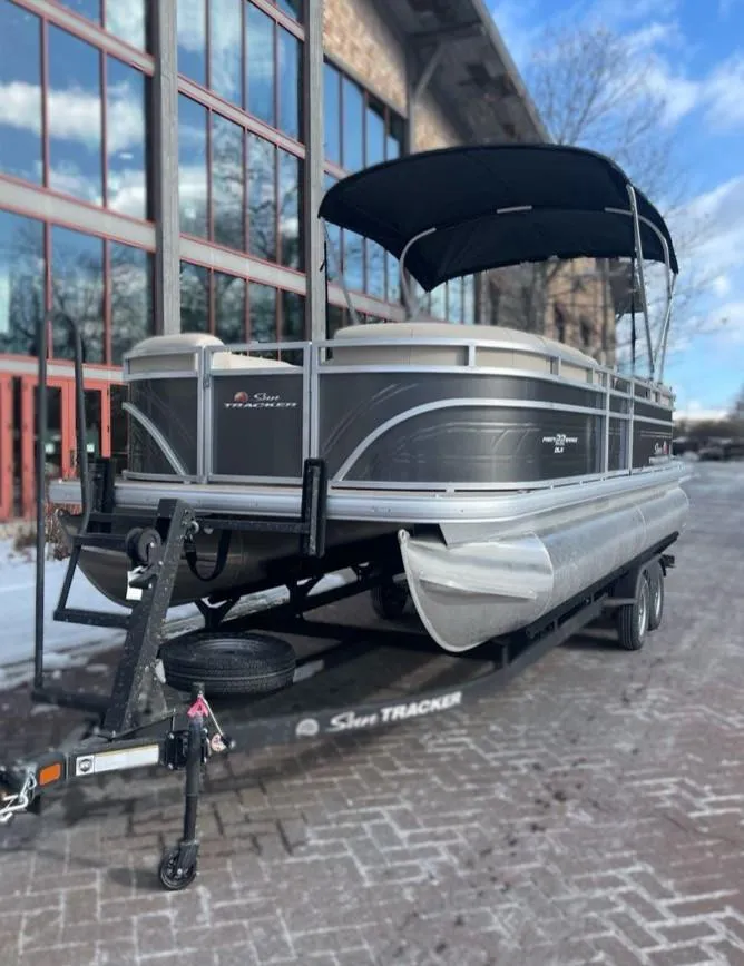 Slide: The Image of 2025 Sun Tracker Party Barge 22 DLX pontoon boat on trailer, parked near a building. - 3