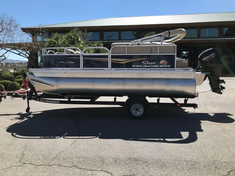 Slide: The Image of 2025 Sun Tracker Bass Buggy 16 XL Select pontoon boat on trailer, parked outdoors. - 2