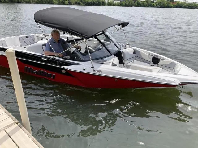 Slide: The Image of 2024 Malibu TXi Mo boat on water near dock, with canopy and driver. - 7