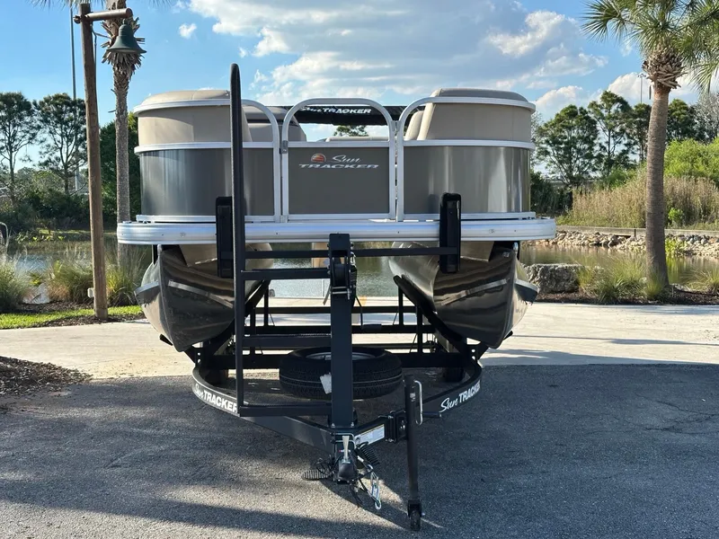 Slide: The Image of 2025 Sun Tracker Party Barge 18 DLX pontoon boat on trailer, parked near a scenic lake. - 2