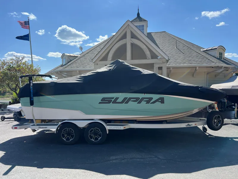 Slide: The Image of 2024 Supra SL boat covered on trailer, parked near a building with flags. - 6