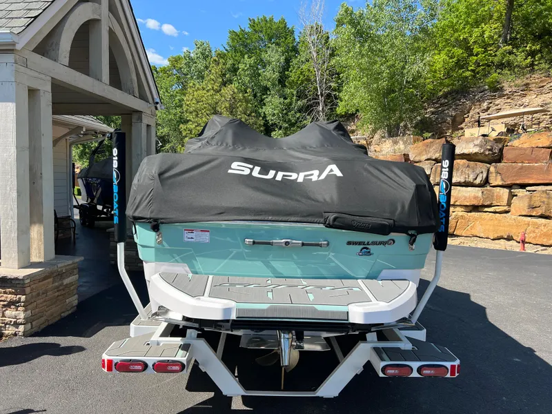 Slide: The Image of 2024 Supra SL boat with cover, parked outdoors, surrounded by trees and stone wall. - 7