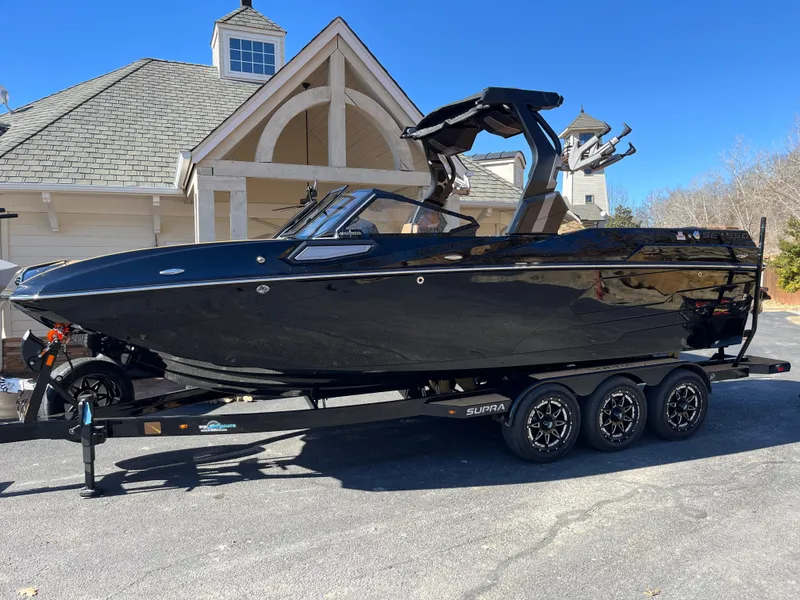 Slide: The Image of 2025 Supra SE boat in black on a trailer, parked near a house. - 0