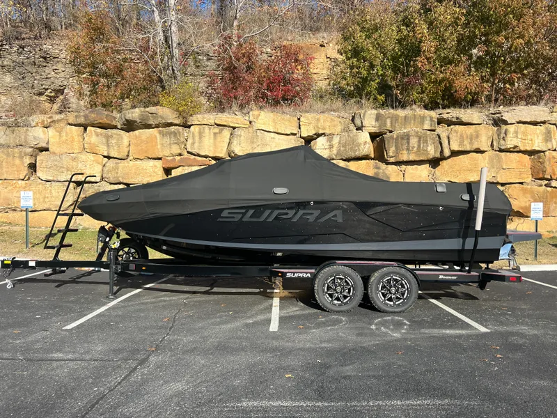 Slide: The Image of 2022 Supra SE boat covered on trailer, parked in front of stone wall. - 22