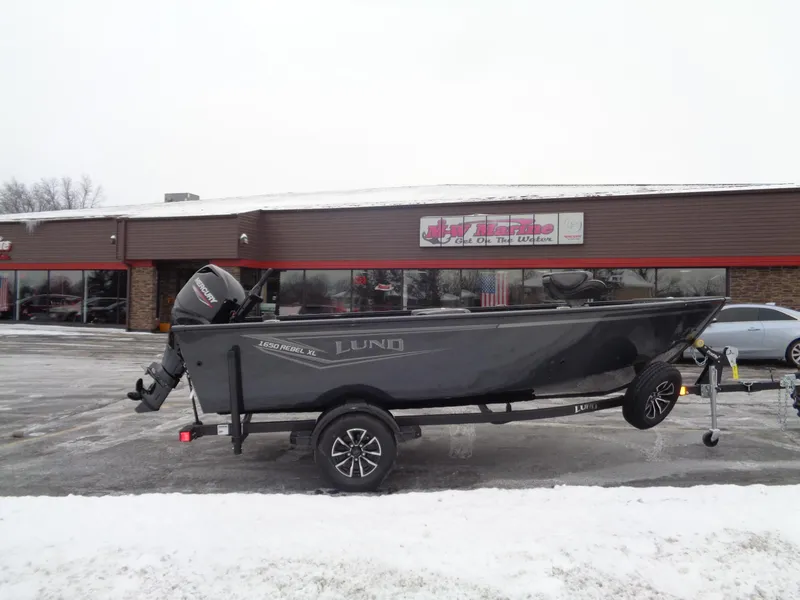 The Image of 2025 Lund 1650 Rebel XL Tiller boat on trailer, parked outside a dealership in snowy conditions. - 0