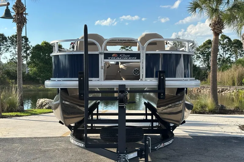 Slide: The Image of 2025 Sun Tracker Bass Buggy 18 DLX pontoon boat on trailer, parked near a scenic lake. - 2