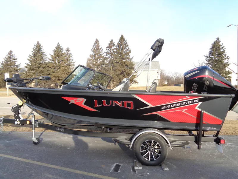 Slide: The Image of 2025 Lund 1875 Crossover XS boat on trailer, black and red design, parked outdoors. - 11