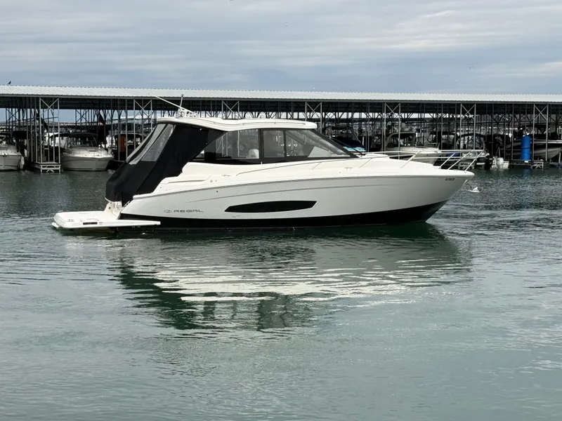 The Image of 2023 Regal 360 Grande Coupe yacht on calm water near a marina. - 0