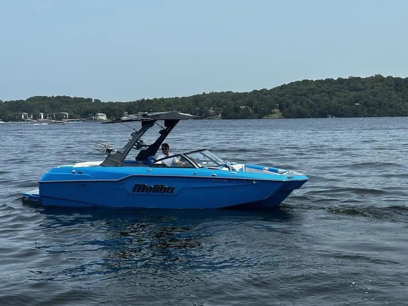 Slide: The Image of Blue 2021 Malibu M220 boat cruising on a lake with forested shoreline. - 2