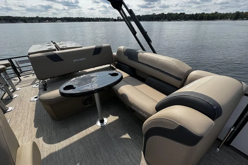 Slide: The Image of 2023 Sylvan L5 DLZ Bar pontoon boat interior with plush seating and table on a lake. - 35