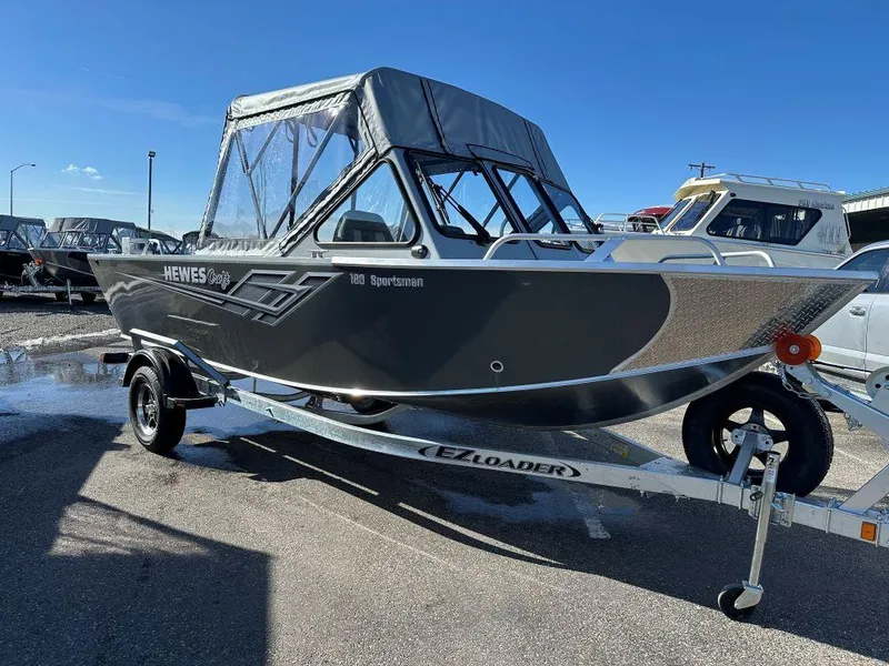The Image of 2025 Hewescraft 180 Sportsman boat on trailer, parked outdoors under clear blue sky. - 0
