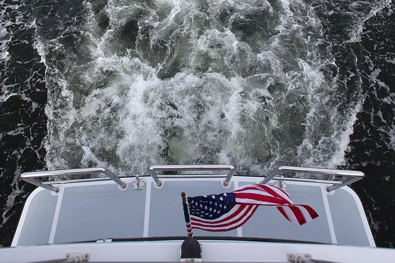 Slide: The Image of Helmsman Trawlers 38E Pilothouse 2026, rear view with American flag, cruising through water. - 16