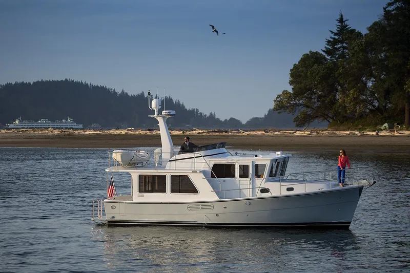 Slide: The Image of 2026 Helmsman Trawlers 38E Pilothouse cruising on calm waters near a forested shoreline. - 2
