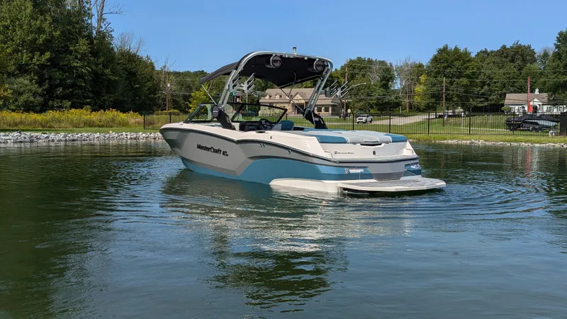 Slide: The Image of 2025 MasterCraft NXT23 boat on a calm lake, surrounded by trees and houses. - 2