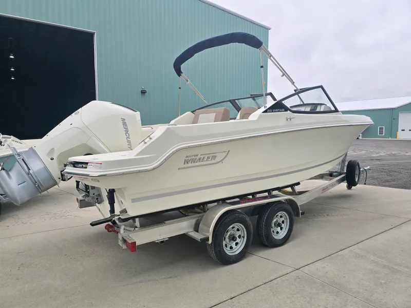 Slide: The Image of 2025 Boston Whaler 210 Vantage boat on trailer, parked outside a large green building. - 5