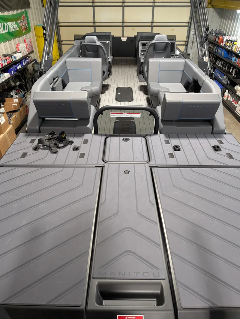 Slide: The Image of 2025 Manitou Explore Dual Engine boat interior with modern seating and storage in a garage setting. - 9