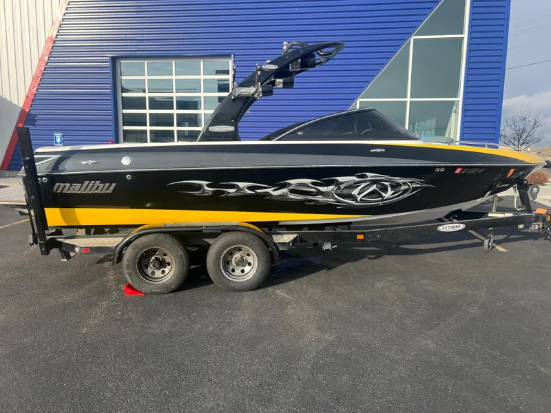 Slide: The Image of 2007 Malibu WVLX boat on trailer, black and yellow design, parked near blue building. - 2