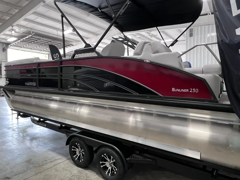 Slide: The Image of 2024 Harris Sunliner 250 pontoon boat with red and black design on a trailer in a showroom. - 3