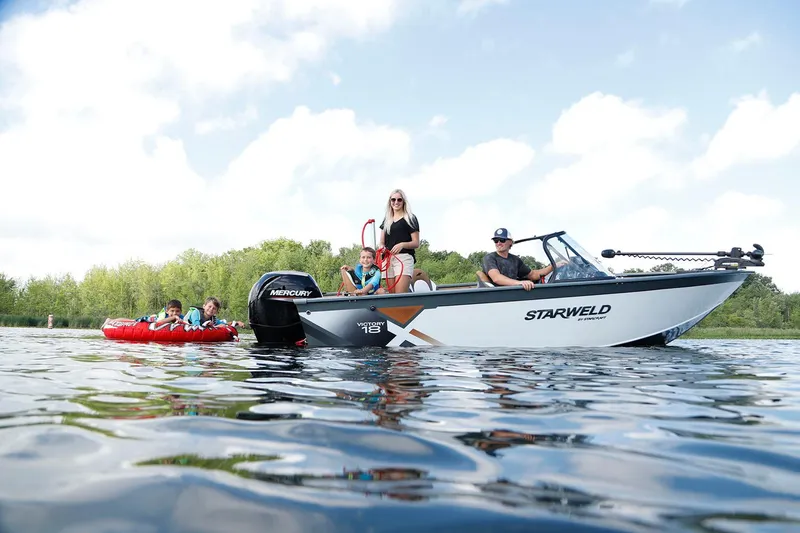 Slide: The Image of 2024 Starweld Victory 18 DC boat on lake with family tubing, clear sky, and lush greenery. - 5