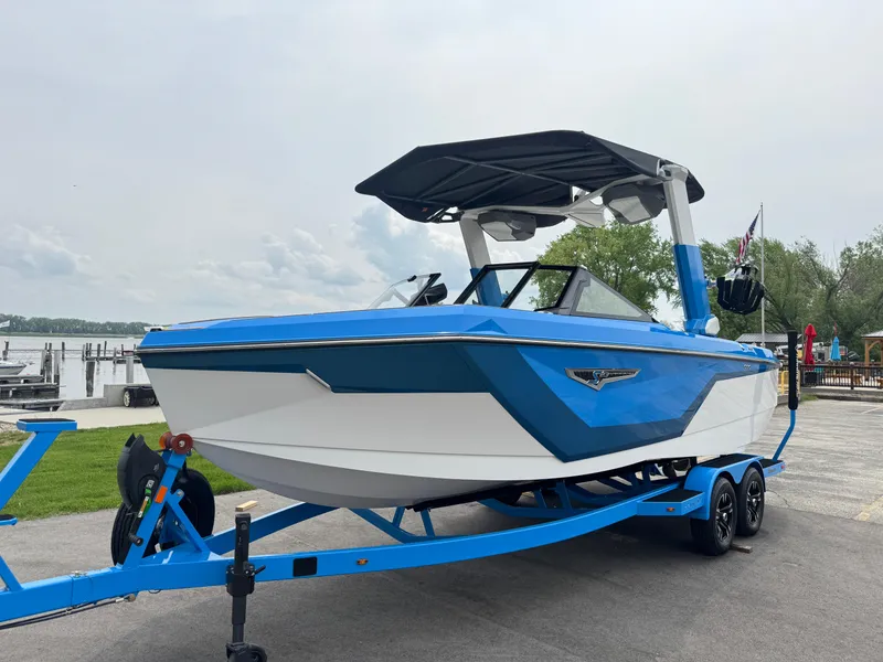 Slide: The Image of 2025 Nautique Super Air Nautique S23 boat in blue and white on a trailer by the water. - 2