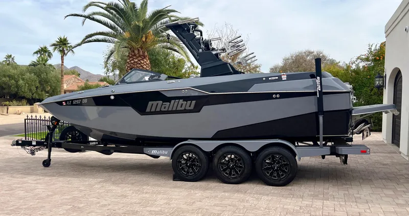 Slide: The Image of 2021 Malibu M240 boat on trailer, parked outdoors with palm trees in the background. - 0