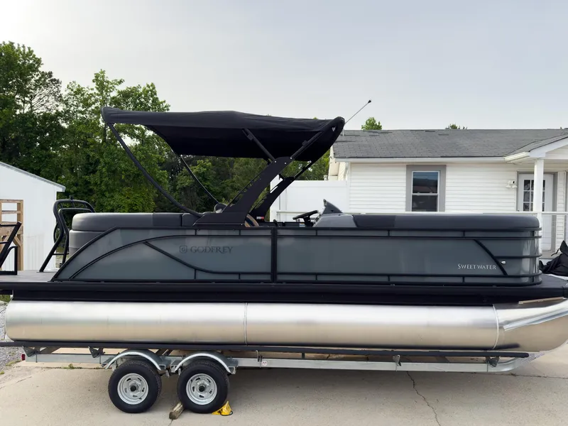 Slide: The Image of 2025 Godfrey Sweetwater 22' Split Bench Tritoon on trailer, parked near a house. - 2