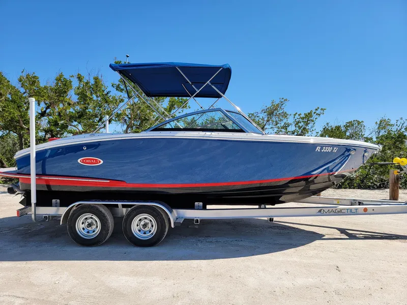 Slide: The Image of 2021 Cobalt CS22 boat on trailer, blue hull, red stripe, sunny day, trees in background. - 12