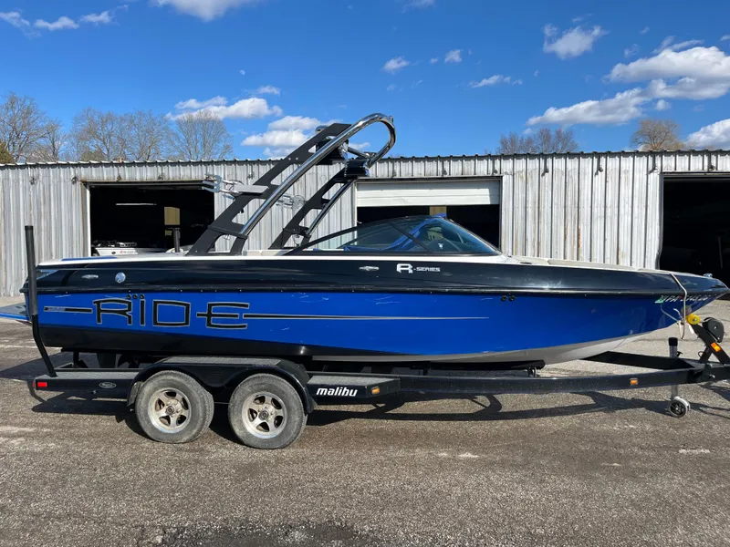 Slide: The Image of 2010 Malibu 21 vRide boat on trailer, blue and black design, parked outdoors. - 0