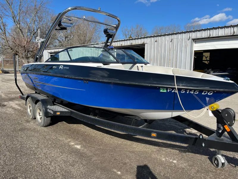Slide: The Image of 2010 Malibu 21 vRide boat on trailer, blue and black design, parked outdoors. - 2