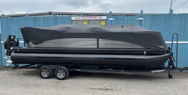 Slide: The Image of 2024 Regency 250 LE3 pontoon boat covered on trailer, parked by blue fence. - 47