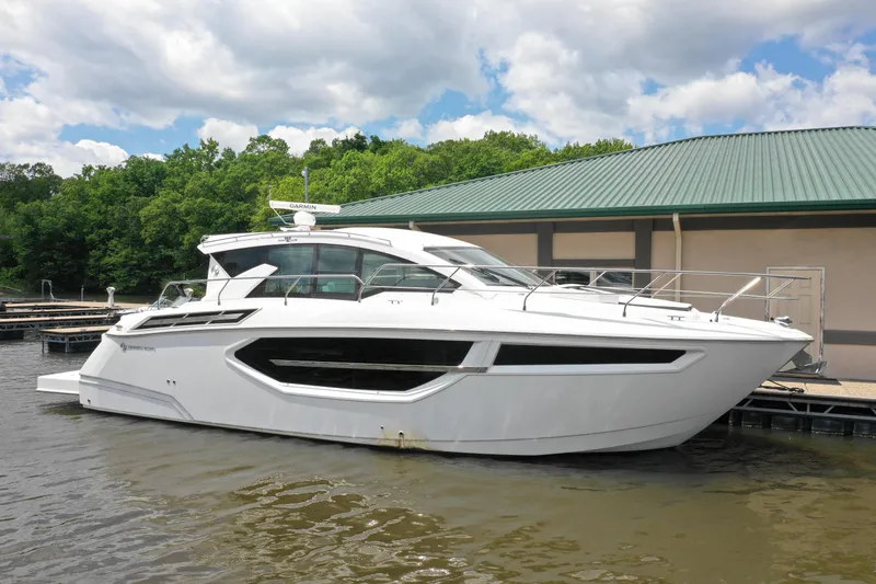Slide: The Image of 2019 Cruisers Yachts 42 Cantius docked by a green-roofed building, surrounded by trees. - 1