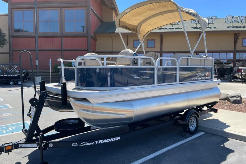 Slide: The Image of 2025 Sun Tracker Bass Buggy 16 XL Select pontoon boat on trailer, parked outdoors. - 2