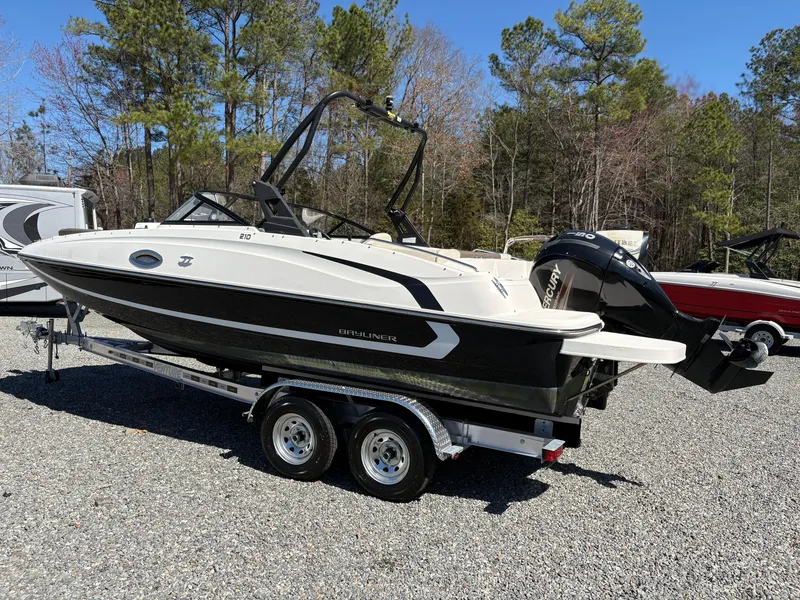 Slide: The Image of 2019 Bayliner 210 Deck Boat on trailer, parked outdoors with trees in the background. - 2