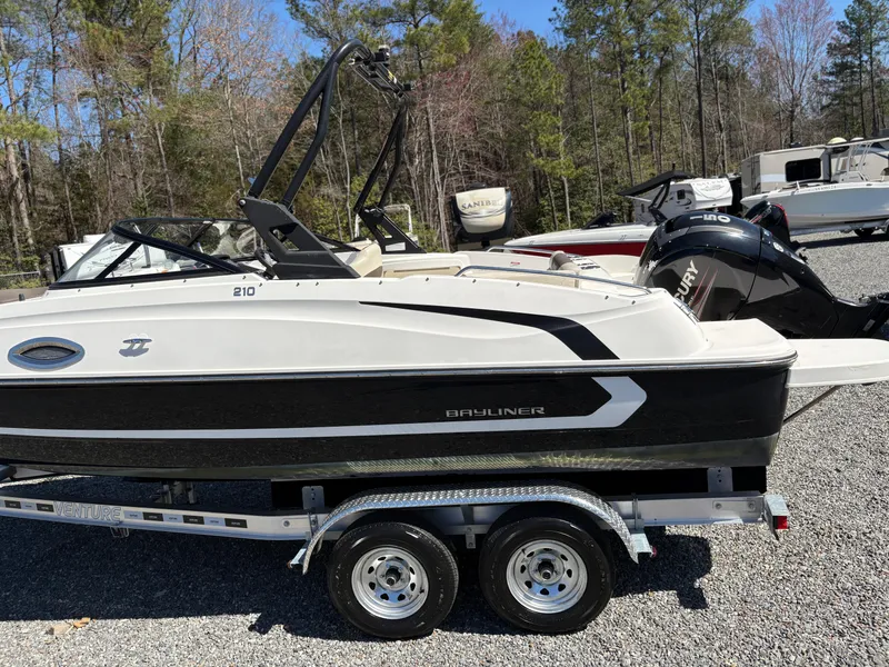 Slide: The Image of 2019 Bayliner 210 Deck Boat on trailer, parked outdoors with trees in background. - 6