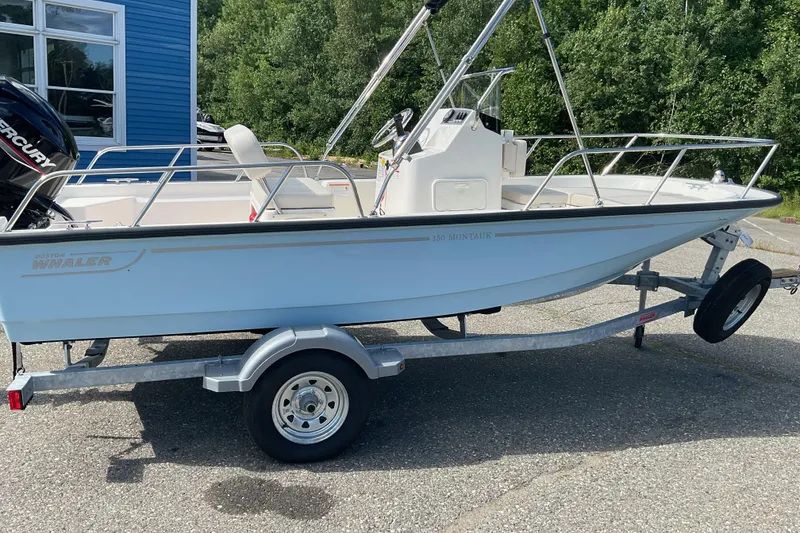 Slide: The Image of 2025 Boston Whaler 150 Montauk boat on trailer, parked outdoors near blue building. - 7